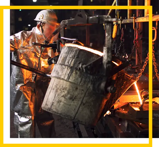 A worker in protective gear pours molten metal from a large crucible into a mold at an industrial foundry, with sparks and glowing liquid metal visible.
