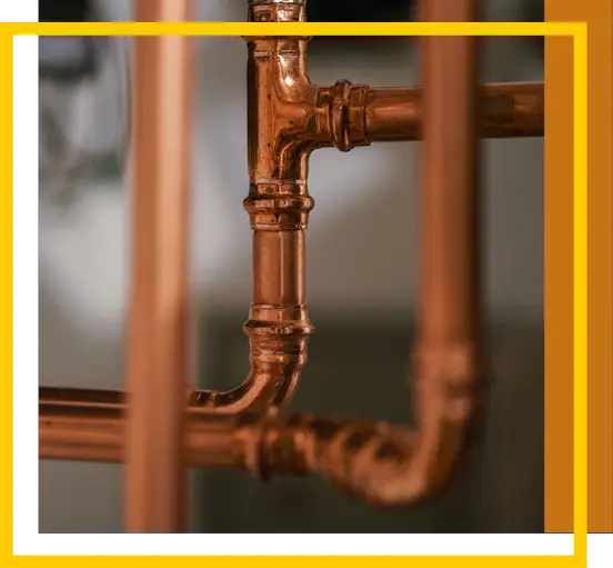 Close-up of shiny copper pipes forming a T-junction, with part of a yellow and white geometric border overlaying the image. The background is blurred, highlighting the metallic texture of the pipes.