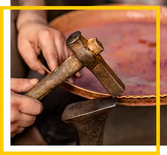 A person uses a hammer and anvil to shape the edge of a round copper plate, working with their hands to create a handcrafted metal object.