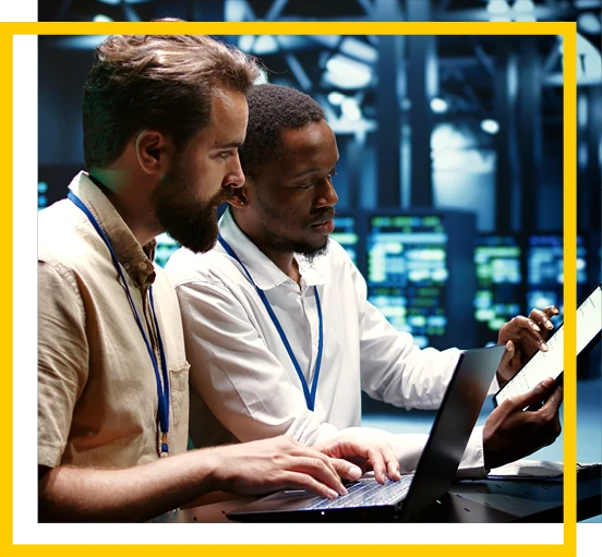 Two men wearing lanyards work together at a laptop in a modern, high-tech office with screens in the background. One types while the other holds a tablet and points at the screen.