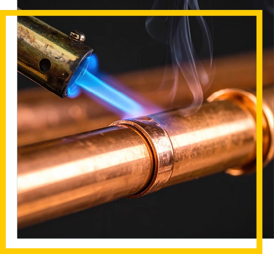 A blue flame from a torch is being used to solder or weld a copper pipe, with smoke rising from the heated joint. The image is bordered by white and yellow lines.