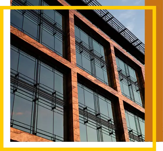 Close-up view of a modern building facade with large reflective glass windows and brown stone framing, photographed from a low angle with a blue sky visible; yellow and white graphic borders frame the image.