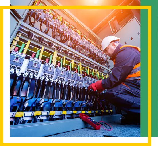 An electrician wearing a hard hat and safety vest works on an open electrical panel filled with wires and switches, using tools and equipment. Bright lighting illuminates the workspace.