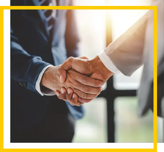 Two people in business suits shaking hands, suggesting a successful agreement or partnership. Sunlight streams in through a window in the blurred background.