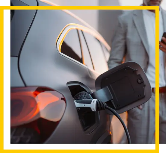 A close-up of an electric vehicle being charged, with a charging cable plugged in. A person in a suit stands in the background, partially visible, holding a phone. The scene is bordered by a yellow frame.