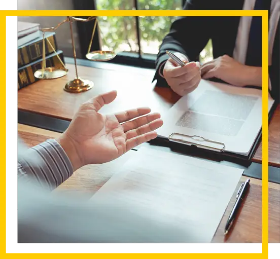 Two people sit at a desk with legal documents, one holding a pen and the other gesturing with an open hand; a scale of justice and books are visible in the background.