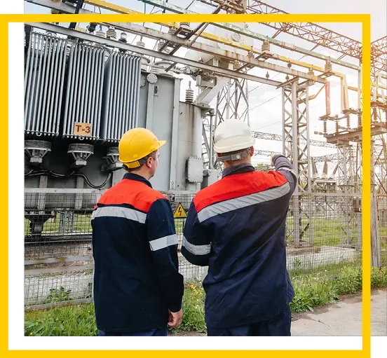Two workers in safety gear and helmets stand outside, with one pointing towards electrical equipment at a power substation. There are metal structures, transformers, and wires visible in the background.