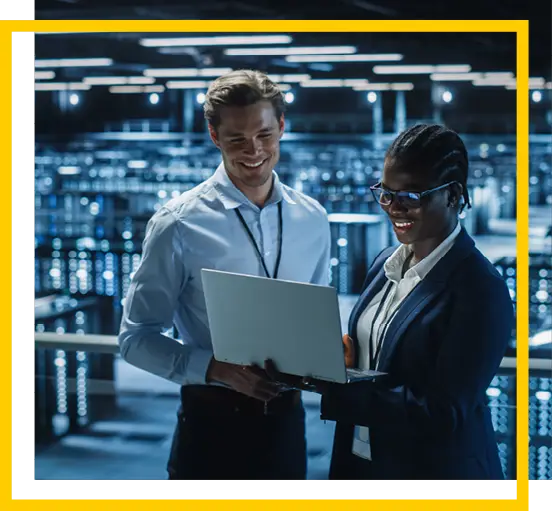 Two professionals, one man and one woman, standing together in a modern data center, smiling and looking at a laptop. Rows of servers and blue lighting are visible in the background.