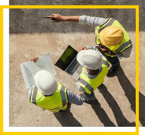 Three construction workers wearing safety vests and helmets review blueprints and a laptop at a job site, with one worker pointing towards something in the distance.