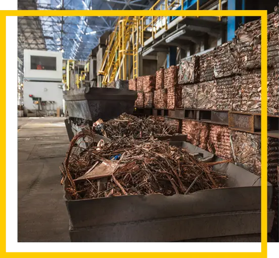 Large bins filled with metal scrap and neatly stacked metal bundles are inside an industrial warehouse with high ceilings and yellow railings.