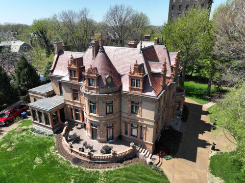 Aerial view of a large, historic mansion with ornate architecture, reddish-brown roof, and beige brick walls, surrounded by trees and a manicured lawn on a sunny day.