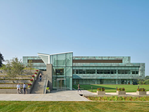A modern glass building with multiple stories features an angular entrance, surrounded by green lawns and tiered stone steps with planters. People walk and sit in the landscaped outdoor area under a clear blue sky.