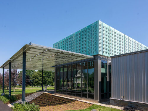 Modern building with large glass windows, an overhanging metal canopy, and a turquoise patterned upper structure. There are trees and grass around the building under a clear blue sky.