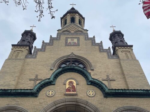 A brick church facade with arched windows, religious icons, and three towers topped with crosses, viewed from below against a cloudy sky. An American flag is visible on the right.