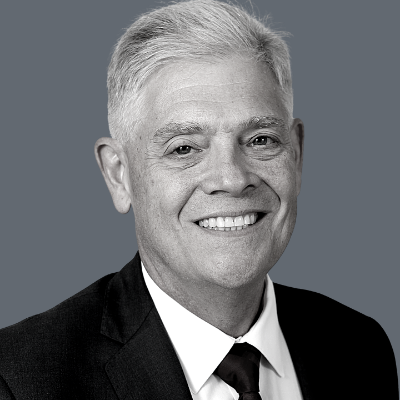 A smiling older man with short gray hair wearing a dark suit, white shirt, and dark tie, posed against a plain gray background.