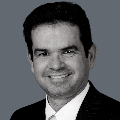 A man with dark hair smiles at the camera, wearing a dark pinstripe suit, white shirt, and tie, in front of a solid gray background.