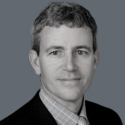 A man with short gray hair wearing a checked shirt, suit jacket, and tie, looking at the camera with a neutral expression against a plain gray background.