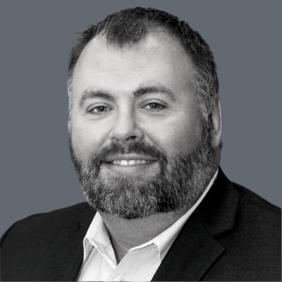 A bearded man wearing a suit jacket and collared shirt smiles at the camera against a plain gray background.