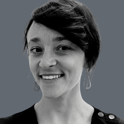 A woman with short dark hair, wearing drop earrings and a dark top with button details, smiles at the camera against a plain gray background.