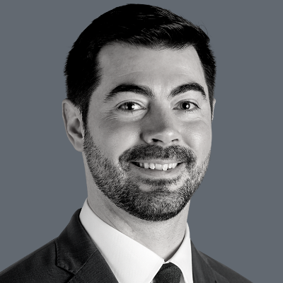 A man with short dark hair and a beard, wearing a suit, white shirt, and tie, smiling at the camera against a plain gray background.