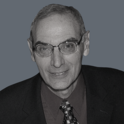 An older man wearing glasses, a dark suit jacket, a maroon shirt, and a patterned tie, smiling at the camera against a plain gray background.