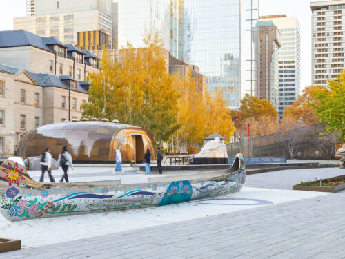 A large, colorful canoe sculpture sits in a modern city plaza with people walking by, surrounded by autumn trees, historic buildings, and glass skyscrapers in the background.