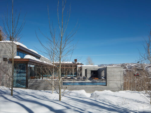 A modern, flat-roofed house with large glass windows sits amid snow-covered ground and leafless trees, with mountains visible in the background under a clear blue sky.