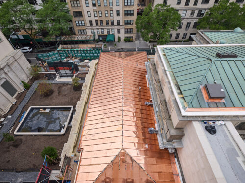 Aerial view looking down at a building with a shiny copper roof, surrounded by other tall buildings, green trees, and a courtyard with soil and planting areas. City street and sidewalks are visible in the background.