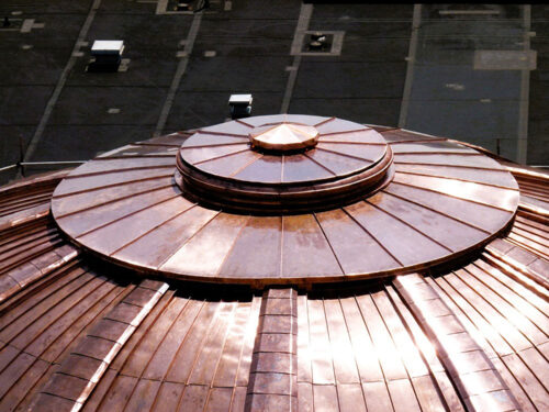 Close-up view of a shiny, segmented copper dome roof with circular, tiered layers reflecting sunlight; dark, flat rooftop and some vents in the background.