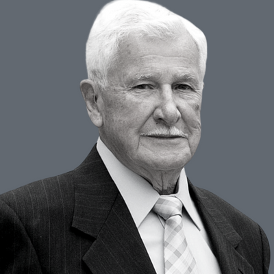 An elderly man with white hair and a mustache, wearing a dark suit, light shirt, and patterned tie, stands against a plain gray background.