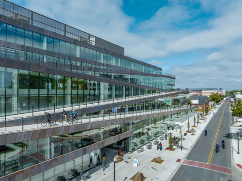 A modern glass building with sloped walkways, where people walk and ride bikes. The structure lines a street with cars, trees, and pedestrians under a partly cloudy sky.