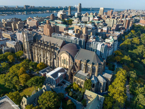 Aerial view of a large Gothic-style cathedral surrounded by trees and city buildings, with a river visible in the background under a clear sky.