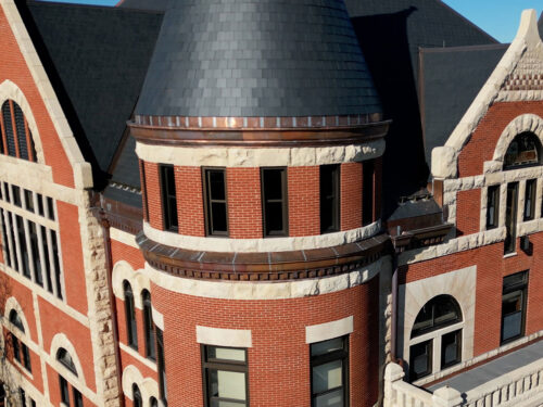 A close-up view of a historic red brick building with arched windows, ornate stone trim, and a slate turret roof under a clear blue sky.