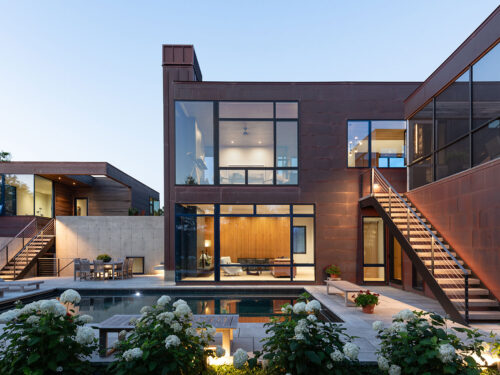 A modern two-story house with large glass windows, an outdoor staircase, a swimming pool, lounge chairs, and landscaped greenery in the foreground, all under a clear sky at dusk.