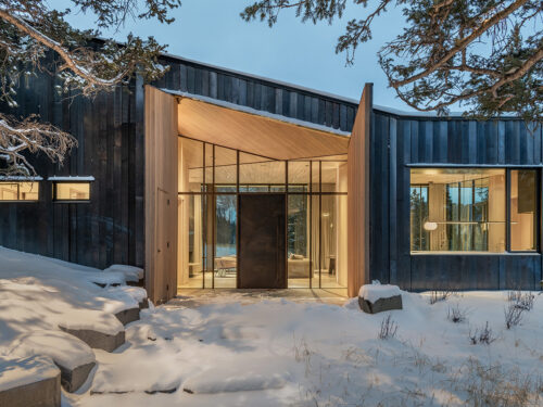 Modern house with black wood siding and large glass entrance, surrounded by snow-covered ground and trees at dusk; warm light glows from the interior windows.