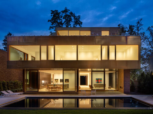 Modern two-story house with large glass windows, warmly lit interior, and a metal exterior. The building overlooks a rectangular pool, surrounded by a patio with lounge chairs, set against a twilight sky and trees.