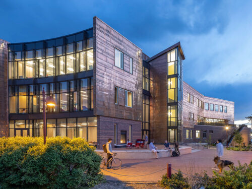 A modern, multi-story building with large glass windows is lit up at dusk. People are walking and biking on a brick pathway outside, and greenery surrounds the area.
