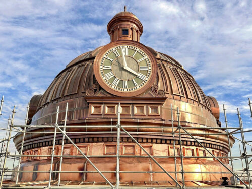 A large copper dome with a clock face is surrounded by scaffolding under a partly cloudy sky, indicating restoration or maintenance work.