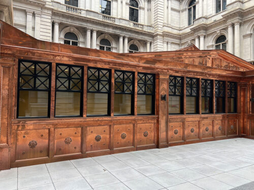 A shiny copper-clad building facade with black-framed windows and ornate detailing stands in a paved courtyard of a grand, historic stone building.