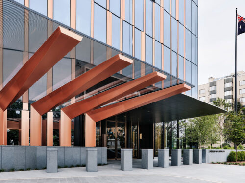 Modern building entrance with large copper beams extending outward above glass doors; reflective glass facade; landscaped greenery and flagpole with Australian flag visible on the right.