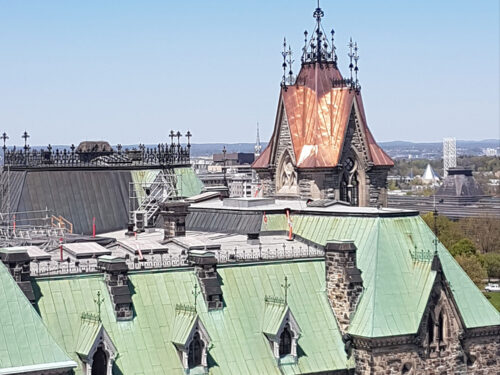 A historic stone building with a green copper roof and an ornate, pointed tower topped with a red-brown spire, set against a clear blue sky and distant landscape.