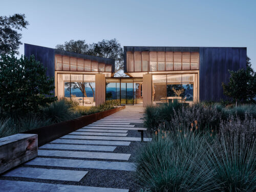A modern house with large glass windows and metal siding, surrounded by lush landscaping and trees, features a stone pathway leading to the entrance at dusk with warm interior lighting visible.